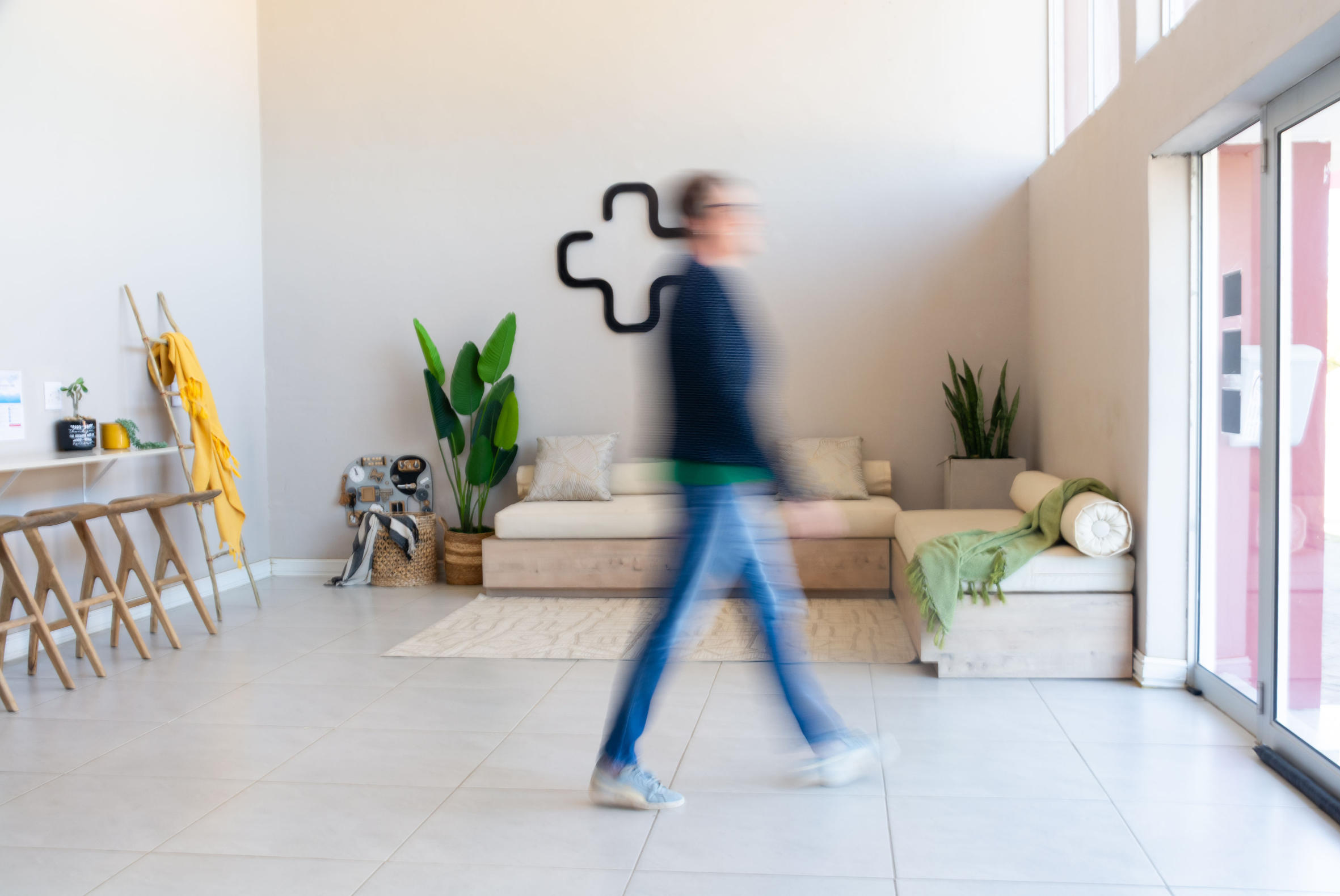 Blurred person walking in a bright modern living room with beige sofas, green plants, wooden stools, and a yellow blanket on a ladder.