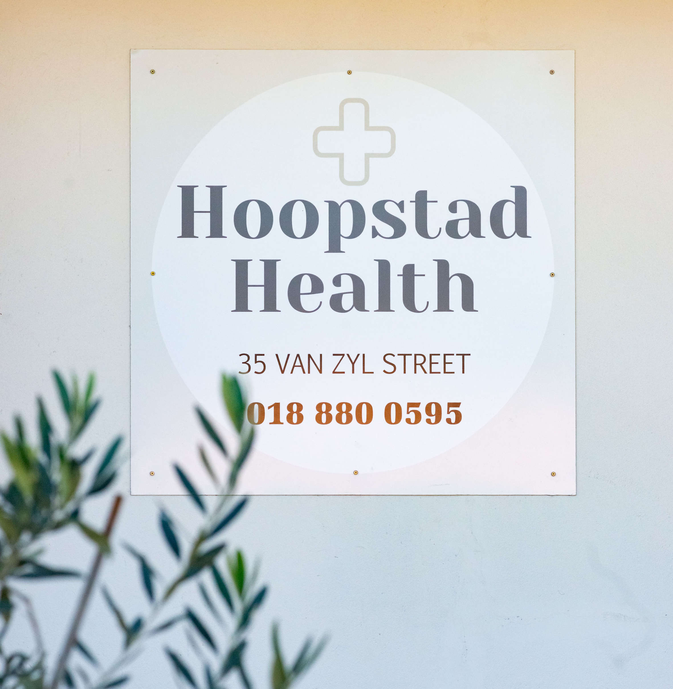 White sign on wall with a medical cross, displaying 'Hoopstad Health', address '35 Van Zyl Street', and phone number '018 880 0595'.