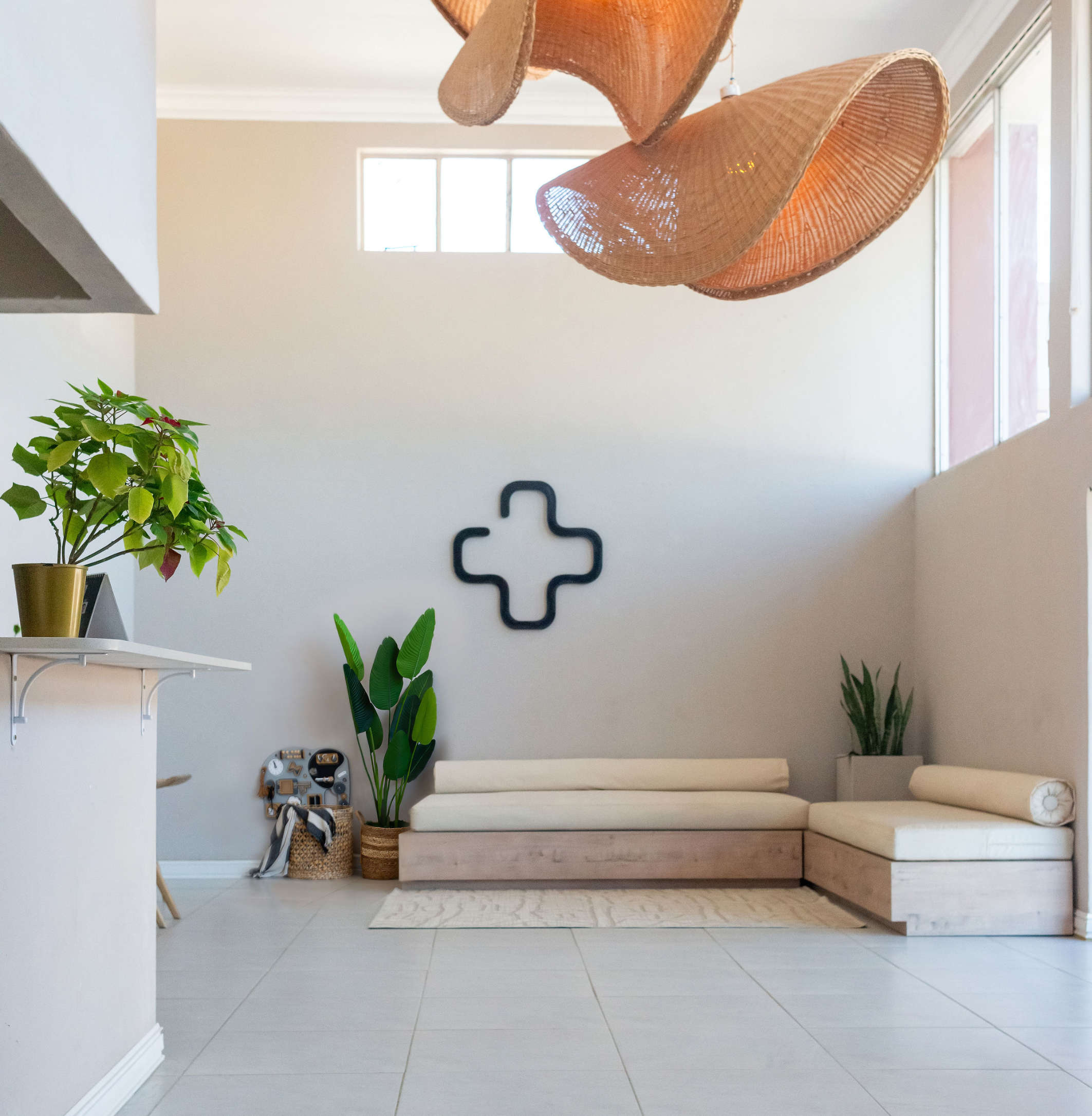 Minimalist waiting area with beige cushioned bench seating, green plants, and modern cross-shaped wall art.