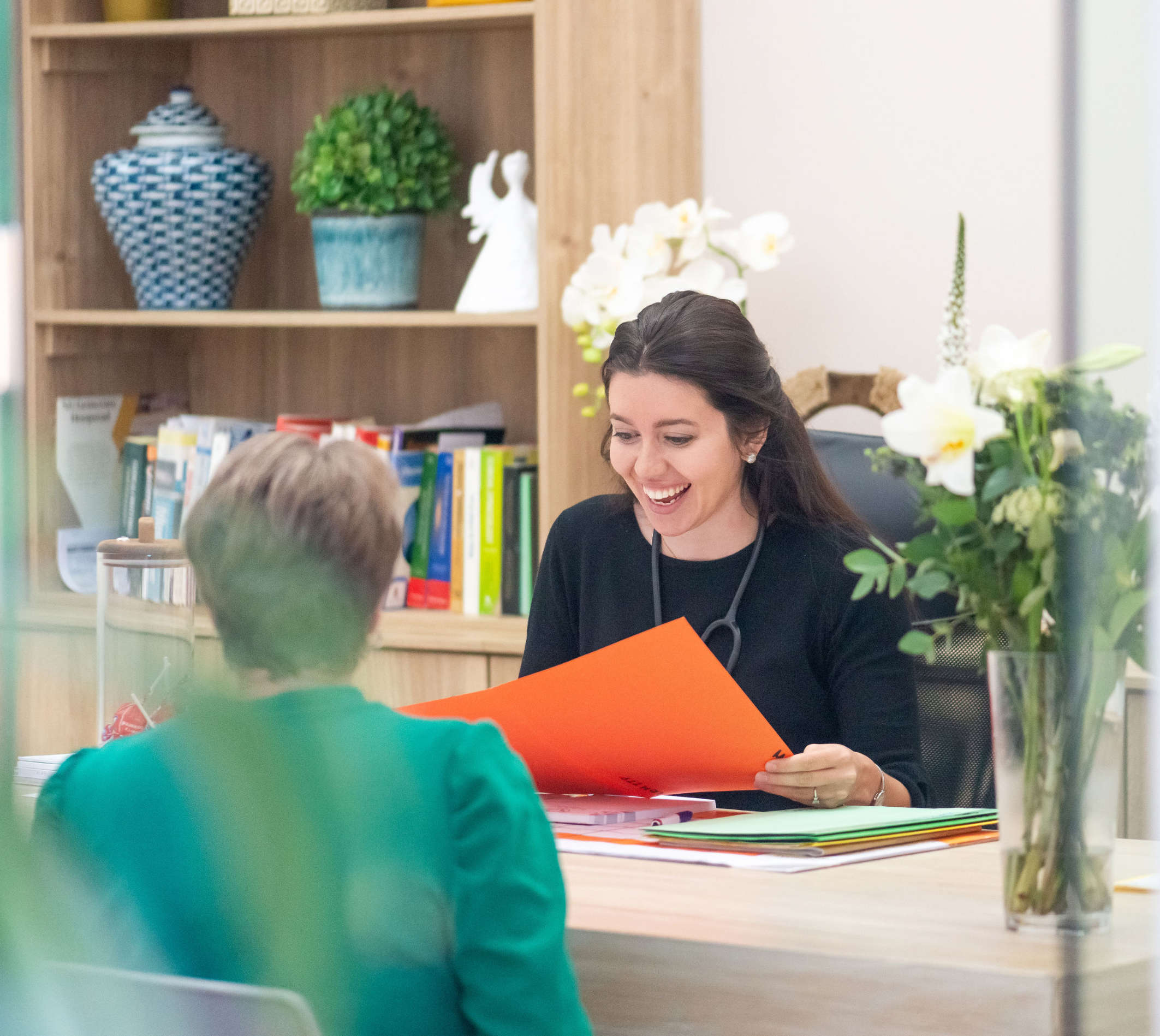 Smiling female doctor reviewing documents with a patient in a bright office with bookshelves and flowers.